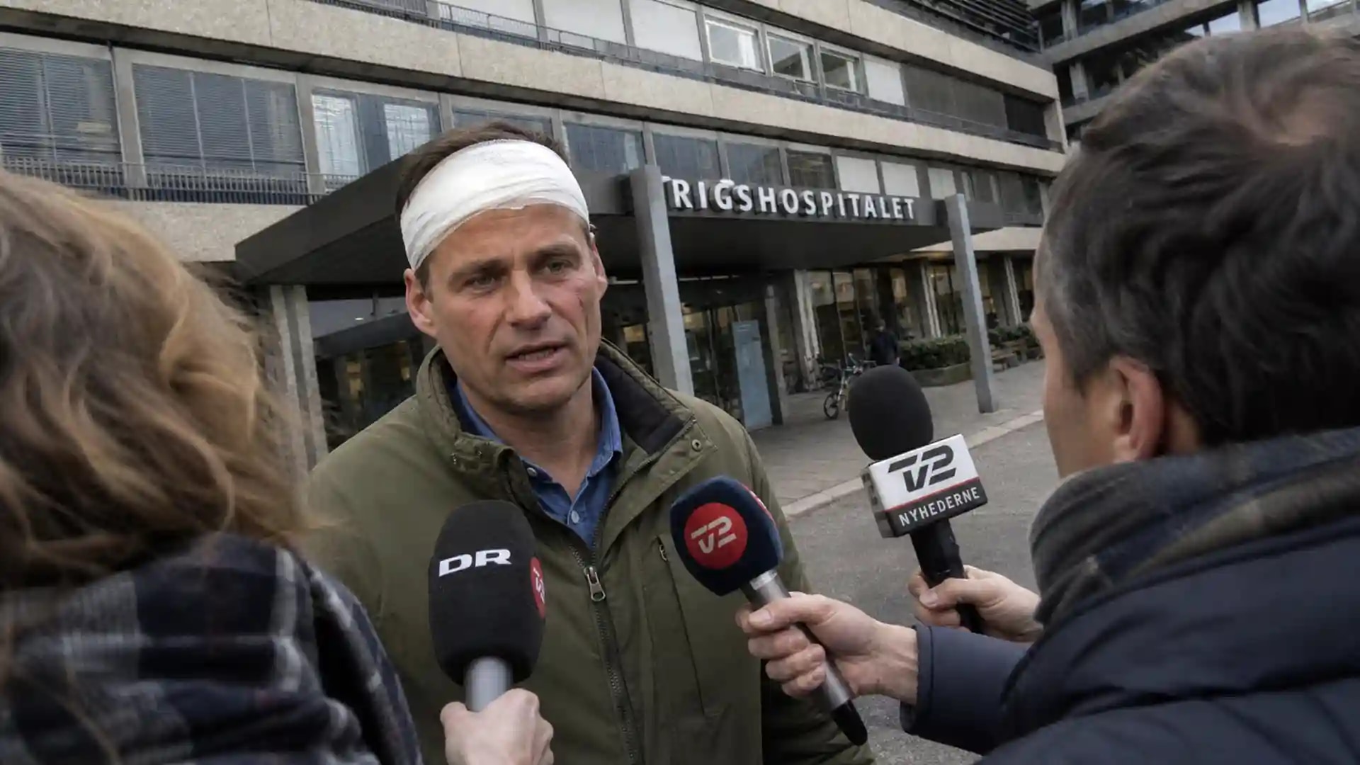 Kim Fournais giver en erklæring til pressen uden for Gentofte Hospital med bandage om hovedet, omringet af journalister og mikrofoner.