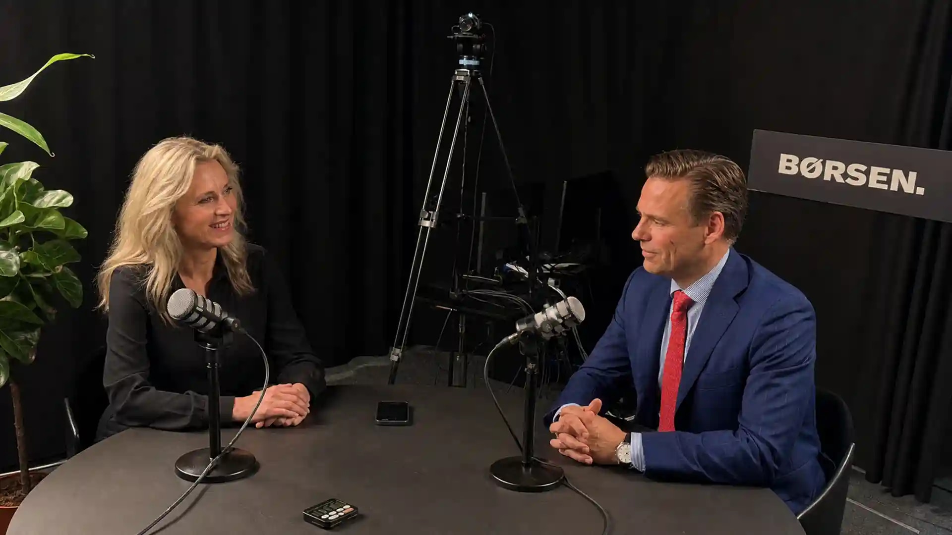 Kim Fournais og værten Tina Riising i Børsens podcaststudie under optagelsen af „Sådan investerer jeg“, mikrofoner og podcastlogo mellem dem.